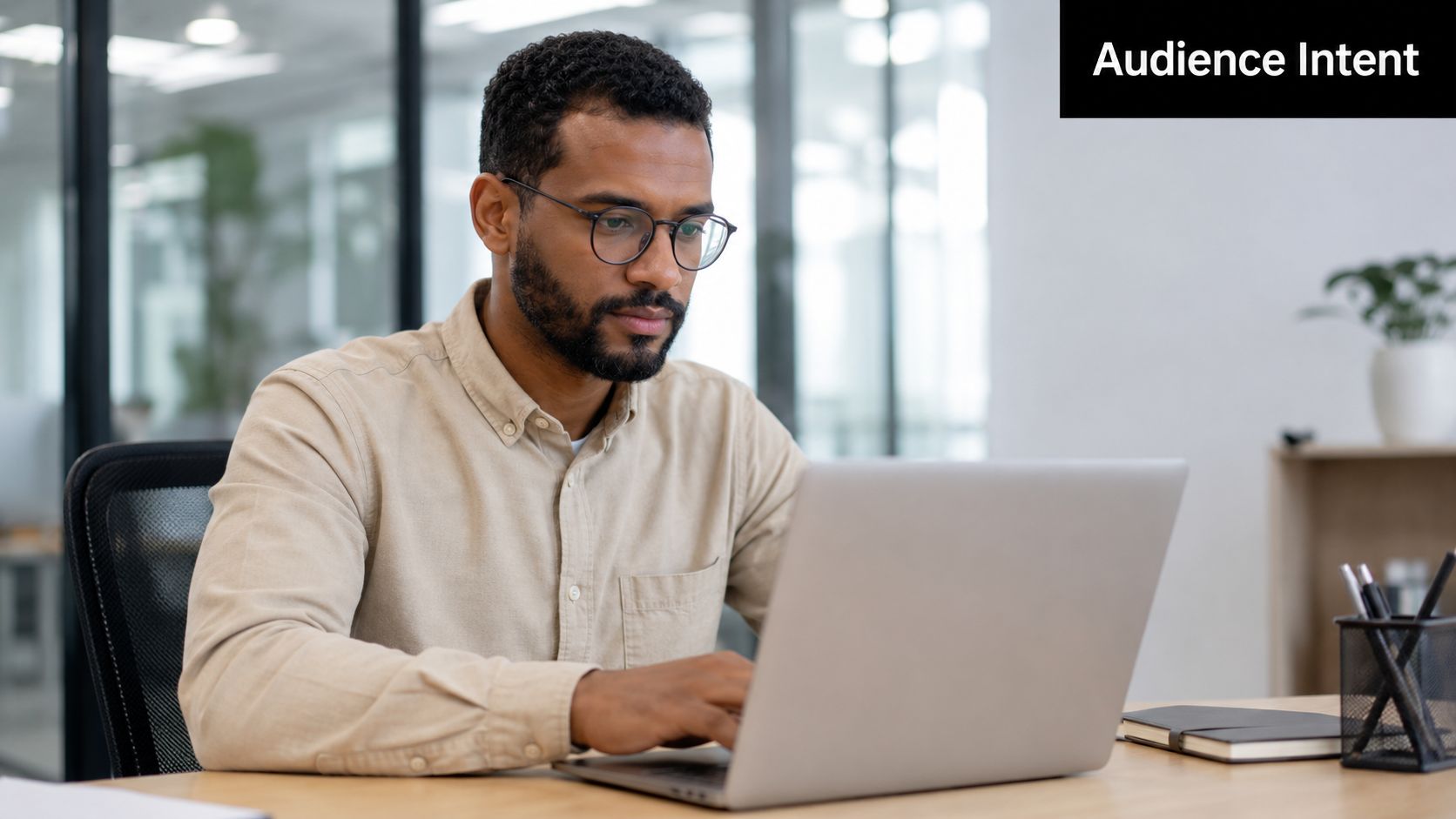 A professional man with glasses working on his laptop in a bright modern office setting.